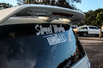 A rear view of a vehicle featuring a large spoiler and a decal on the back window with the text 'Street Works'. In the background, another vehicle, possibly a pickup truck, is parked on the side of the road with trees and greenery visible.