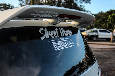 A rear view of a vehicle featuring a large spoiler and a decal on the back window with the text 'Street Works'. In the background, another vehicle, possibly a pickup truck, is parked on the side of the road with trees and greenery visible.