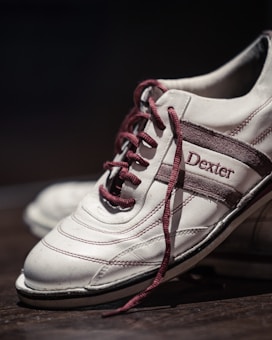 A close-up of a white athletic shoe with burgundy accents and laces. The shoe displays the brand name 'Dexter' stitched in burgundy on the side. The focus is on the shoe's intricate stitching and elegant design with a softly blurred background.
