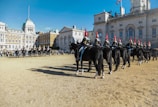 A line of mounted soldiers in uniform, wearing red plumed helmets, is positioned in a historical courtyard. These soldiers are riding black horses that stand in neat ranks. The backdrop includes a large, classical building with multiple arched windows, and a crowd of spectators lines the perimeter of the open space.