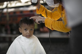 A young child with a solemn expression is receiving a haircut. The barber, wearing a yellow shirt and a face mask, carefully uses clippers to trim the child's hair. The child is draped in a white cape, and the setting appears to be informal, possibly outdoors or in a rustic environment.