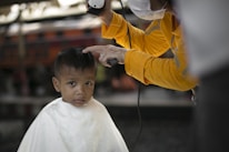 A young child with a solemn expression is receiving a haircut. The barber, wearing a yellow shirt and a face mask, carefully uses clippers to trim the child's hair. The child is draped in a white cape, and the setting appears to be informal, possibly outdoors or in a rustic environment.