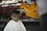 A young child with a solemn expression is receiving a haircut. The barber, wearing a yellow shirt and a face mask, carefully uses clippers to trim the child's hair. The child is draped in a white cape, and the setting appears to be informal, possibly outdoors or in a rustic environment.