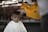 A young child with a solemn expression is receiving a haircut. The barber, wearing a yellow shirt and a face mask, carefully uses clippers to trim the child's hair. The child is draped in a white cape, and the setting appears to be informal, possibly outdoors or in a rustic environment.