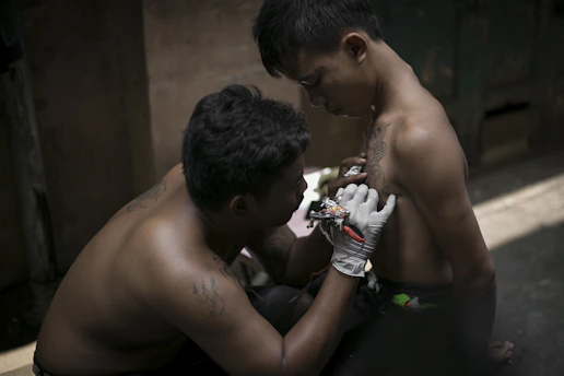A shirtless person is getting a tattoo on their shoulder from another shirtless person in a dimly lit setting. The person doing the tattoo is wearing gloves and using a tattoo machine.