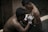 A shirtless person is getting a tattoo on their shoulder from another shirtless person in a dimly lit setting. The person doing the tattoo is wearing gloves and using a tattoo machine.