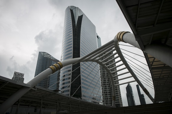 A futuristic urban landscape with a modern architectural design featuring a prominent skyscraper with glass facades. The structure is accentuated by a distinctive white arch with cables and metallic details in the foreground. The sky is overcast, adding a dramatic effect to the scene.