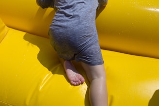 A child wearing a wet grey t-shirt and pants is climbing on a bright yellow inflatable surface. The child's right leg and foot are visible, showing movement and playfulness.