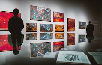 An art gallery features a series of abstract paintings on a grey wall, varying in size and consisting of rich colors like reds, blues, and oranges. Two individuals stand in front of the paintings, each observing the artwork intently. The highly polished floor reflects the paintings and people, creating a mirror effect. On the surface in the foreground, some documents or papers are visible, adding to the gallery's ambiance.