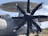 A large military aircraft featuring a prominent propeller and radar dome. The propeller has multiple blades with a sleek, modern design. The aircraft's body is a light gray color with red accents and labeled with cautionary text. The setting appears to be an airfield with a blue sky and some structures visible in the background.