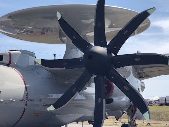 A large military aircraft featuring a prominent propeller and radar dome. The propeller has multiple blades with a sleek, modern design. The aircraft's body is a light gray color with red accents and labeled with cautionary text. The setting appears to be an airfield with a blue sky and some structures visible in the background.