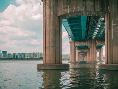 A large concrete bridge extends over a wide body of water. The understructure of the bridge is visible, with multiple supporting columns reflecting in the water. In the background, a skyline of modern apartment buildings stretches across the horizon under a lightly clouded sky.