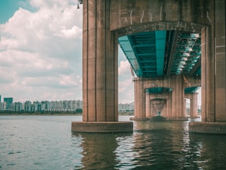 A large concrete bridge extends over a wide body of water. The understructure of the bridge is visible, with multiple supporting columns reflecting in the water. In the background, a skyline of modern apartment buildings stretches across the horizon under a lightly clouded sky.