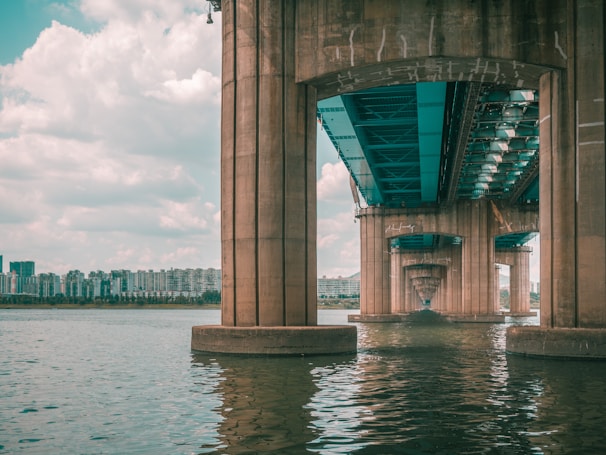 A large concrete bridge extends over a wide body of water. The understructure of the bridge is visible, with multiple supporting columns reflecting in the water. In the background, a skyline of modern apartment buildings stretches across the horizon under a lightly clouded sky.