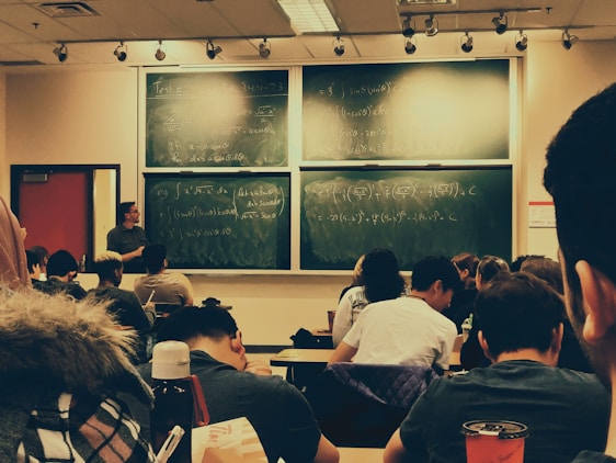 A classroom setting with a group of students seated and facing a large chalkboard filled with mathematical equations and diagrams. A teacher stands at the front, engaging with the students. The room has a warm lighting atmosphere with track lights on the ceiling, and various personal items like water bottles are visible on the desks.