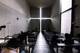 A dimly lit, modern chapel space with concrete walls and a large cross formed by the intersection of light beams at the front. The interior has benches where people sit, some looking at their phones or reading.