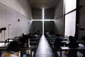 A dimly lit, modern chapel space with concrete walls and a large cross formed by the intersection of light beams at the front. The interior has benches where people sit, some looking at their phones or reading.