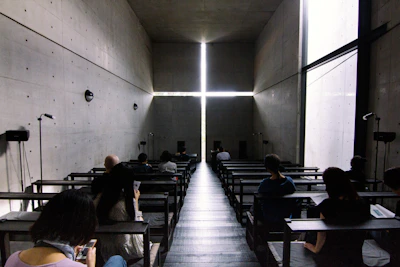 A dimly lit, modern chapel space with concrete walls and a large cross formed by the intersection of light beams at the front. The interior has benches where people sit, some looking at their phones or reading.