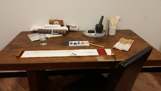 A wooden table is set up with various items related to painting and writing. On the table are scrolls, a book, a cup of water, a palette with watercolor paints, brushes in a holder, a stone ink set, and an unfolded scroll with a brush resting on top. The scene suggests an artistic or calligraphic workspace.