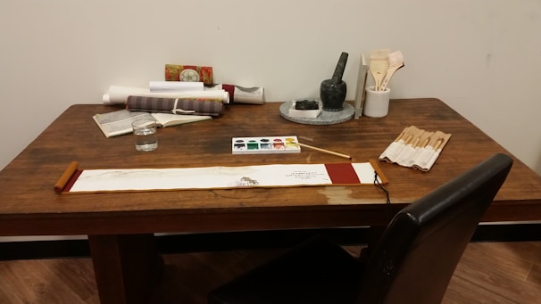 A wooden table is set up with various items related to painting and writing. On the table are scrolls, a book, a cup of water, a palette with watercolor paints, brushes in a holder, a stone ink set, and an unfolded scroll with a brush resting on top. The scene suggests an artistic or calligraphic workspace.