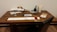 A wooden table is set up with various items related to painting and writing. On the table are scrolls, a book, a cup of water, a palette with watercolor paints, brushes in a holder, a stone ink set, and an unfolded scroll with a brush resting on top. The scene suggests an artistic or calligraphic workspace.