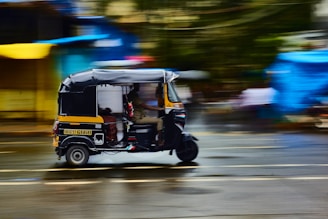 A black and yellow auto rickshaw is in motion, speeding along a wet, blurred city street. The background features colorful blurred shapes, indicating a vivid, bustling urban environment. The street surface reflects light, suggesting recent rainfall.