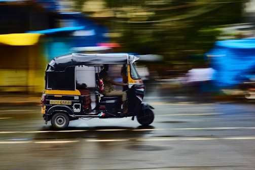 A black and yellow auto rickshaw is in motion, speeding along a wet, blurred city street. The background features colorful blurred shapes, indicating a vivid, bustling urban environment. The street surface reflects light, suggesting recent rainfall.