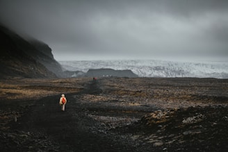 A lone person wearing a bright red backpack walks along a dark, rocky path with a vast, rugged landscape stretching into the distance. The sky is overcast, casting a moody and dramatic atmosphere over the scene. In the distance, a massive glacier is visible, partially shrouded in mist.