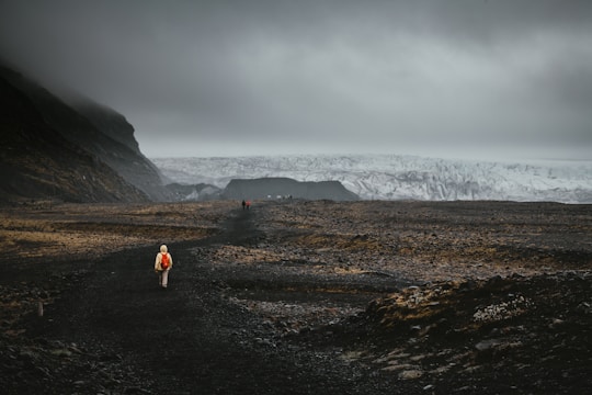 A lone person wearing a bright red backpack walks along a dark, rocky path with a vast, rugged landscape stretching into the distance. The sky is overcast, casting a moody and dramatic atmosphere over the scene. In the distance, a massive glacier is visible, partially shrouded in mist.