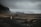 A lone person wearing a bright red backpack walks along a dark, rocky path with a vast, rugged landscape stretching into the distance. The sky is overcast, casting a moody and dramatic atmosphere over the scene. In the distance, a massive glacier is visible, partially shrouded in mist.