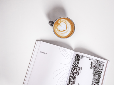 A flat lay featuring a cup of cappuccino with a latte art heart next to an open book titled 'Nothingness'. The book displays abstract black and white artwork with a silhouette on the right page.