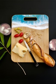 A wooden cutting board with a design resembling a beach shoreline holds sliced bread, Swiss cheese, cherry tomatoes, olives, and pine nuts. A knife rests near the bread. Two glasses with ice are placed on either side, and a green plant stem decorates the left corner.
