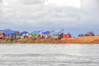 A makeshift settlement with blue and green tarpaulin tents lined along a muddy bank beside a body of water. Several motorcycles are parked nearby, and people are seen near the tents. The sky is cloudy, suggesting an overcast day.