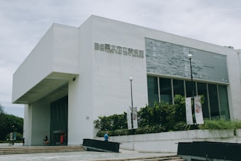 A modern, white building with the words Taipei Fine Arts Museum on the facade. The structure features clean lines and large windows, with lamp posts and banners at the entrance. Trees and shrubs add greenery around the museum.