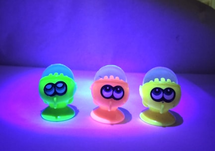 Three brightly colored toy figures with large eyes are illuminated under a UV light. The figures are made of transparent material that makes them glow in neon shades of green, orange, and yellow. They are standing upright on a flat surface with a purple and blue gradient background.