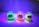 Three brightly colored toy figures with large eyes are illuminated under a UV light. The figures are made of transparent material that makes them glow in neon shades of green, orange, and yellow. They are standing upright on a flat surface with a purple and blue gradient background.
