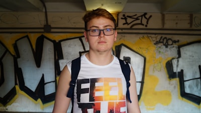 A person wearing glasses and a sleeveless shirt stands in front of a graffiti-covered wall. The shirt has a color gradient design with the words 'FEEL THE' visible. The wall showcases various graffiti art with bold black outlines over a yellow and grayish background.