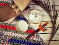 A traditional setting featuring a woven mat with a colorful, geometric-patterned textile in red, blue, and green. On top of the textile are bamboo trays, one containing white flowers. A wooden box, a traditional dagger with a decorative handle, and dried palm leaves are also present, all arranged on wooden planks.