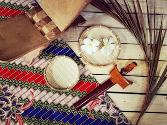 A traditional setting featuring a woven mat with a colorful, geometric-patterned textile in red, blue, and green. On top of the textile are bamboo trays, one containing white flowers. A wooden box, a traditional dagger with a decorative handle, and dried palm leaves are also present, all arranged on wooden planks.