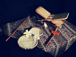A traditional setup featuring a variety of cultural items on a patterned fabric background. In the foreground, there are white flowers placed on circular woven trays. To the side, a wooden object resembling a sword or ceremonial dagger rests. A metal fan-like object with a long handle is also visible, suggesting an artistic or cultural theme.