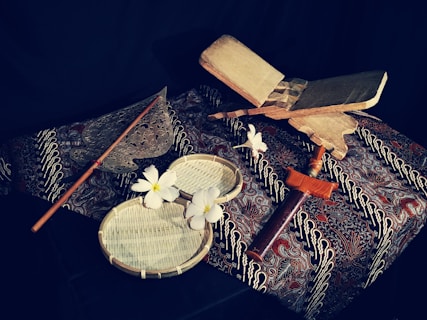 A traditional setup featuring a variety of cultural items on a patterned fabric background. In the foreground, there are white flowers placed on circular woven trays. To the side, a wooden object resembling a sword or ceremonial dagger rests. A metal fan-like object with a long handle is also visible, suggesting an artistic or cultural theme.