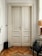 A double panel wooden door with ornate detailing, set in a simple interior with light-colored walls and a parquet floor. To the right of the door stands a modern chair with a glass seat, next to a woven lamp on a windowsill. On the left, there is a black cylindrical wastebasket.