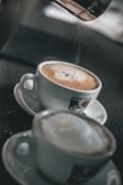 Two cups of coffee are placed on saucers, with a focus on the frothy top of each drink. One cup is being sprinkled with chocolate powder from a container above. The cups are white and feature a logo with the word 'Einstein'. The background is softly blurred, highlighting the focus on the coffee cups.