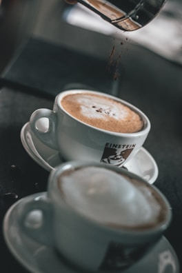 Two cups of coffee are placed on saucers, with a focus on the frothy top of each drink. One cup is being sprinkled with chocolate powder from a container above. The cups are white and feature a logo with the word 'Einstein'. The background is softly blurred, highlighting the focus on the coffee cups.