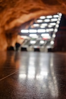 A smooth, reflective floor stretches out towards an abstract structure composed of angled, illuminated panels. The warm, rocky ceiling curves above the scene, suggesting an underground or cave-like environment. The lighting creates a dramatic contrast with the darker surroundings.