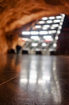 A smooth, reflective floor stretches out towards an abstract structure composed of angled, illuminated panels. The warm, rocky ceiling curves above the scene, suggesting an underground or cave-like environment. The lighting creates a dramatic contrast with the darker surroundings.