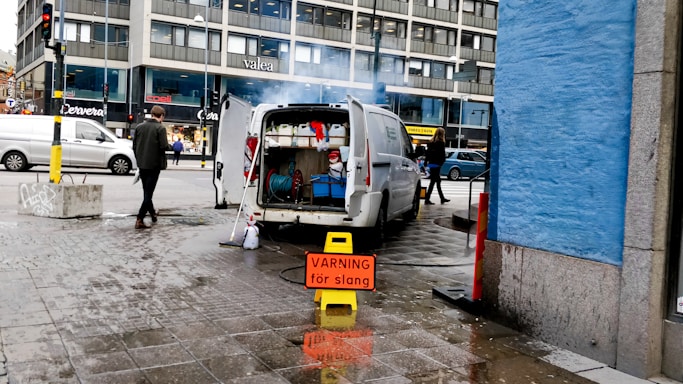 A white van with its rear doors open is parked on an urban street. Inside the van, various cleaning or maintenance equipment is visible, including hoses and buckets. A prominent yellow warning sign with red text in Swedish is placed on the wet pavement near the van. Pedestrians walk along the sidewalk, and a blue-painted building wall is on the right. A large office or commercial building is in the background.