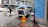 A white van with its rear doors open is parked on an urban street. Inside the van, various cleaning or maintenance equipment is visible, including hoses and buckets. A prominent yellow warning sign with red text in Swedish is placed on the wet pavement near the van. Pedestrians walk along the sidewalk, and a blue-painted building wall is on the right. A large office or commercial building is in the background.