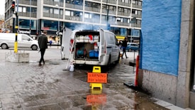 A white van with its rear doors open is parked on an urban street. Inside the van, various cleaning or maintenance equipment is visible, including hoses and buckets. A prominent yellow warning sign with red text in Swedish is placed on the wet pavement near the van. Pedestrians walk along the sidewalk, and a blue-painted building wall is on the right. A large office or commercial building is in the background.