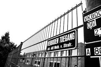 A black and white image of a metal fence with a sign reading 'Verboden Toegang' in Dutch, translating to 'No Trespassing'. The sign references a specific legal article. In the background, part of a building and a tree are visible.